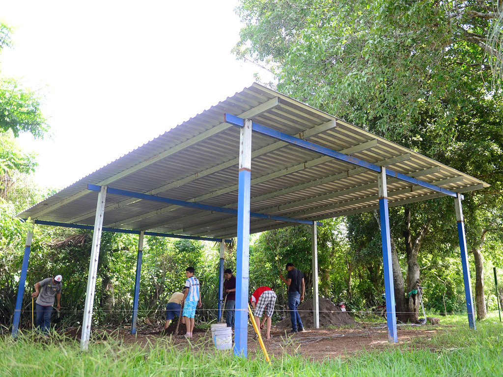 Un grupo de cinco personas, entre ellos jóvenes y adultos, trabajando con palas y baldes en el suelo de tierra debajo de una estructura de techo de lámina recién levantada con postes azules, rodeados de vegetación.
