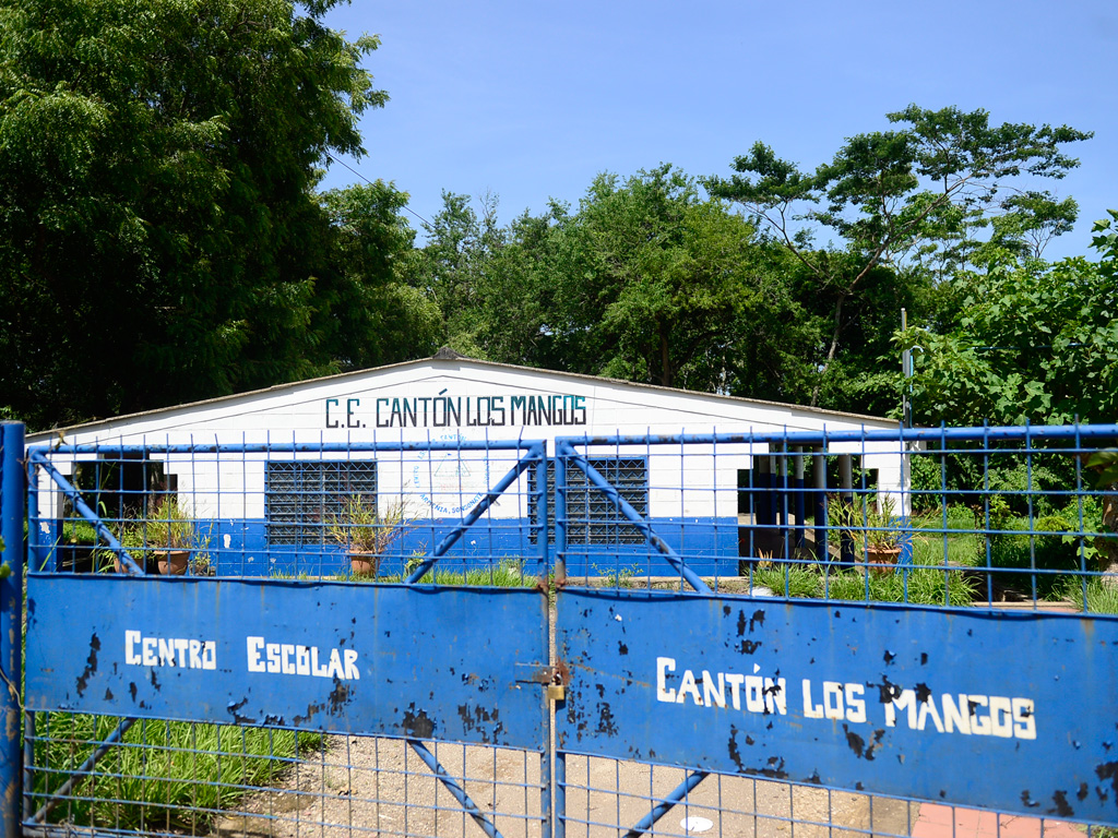Fachada exterior y portón metálico de color azul con letras blancas que dicen "C.E. Cantón Los Mangos" en Armenia, cerrado y rodeado de vegetación crecida bajo un cielo despejado.