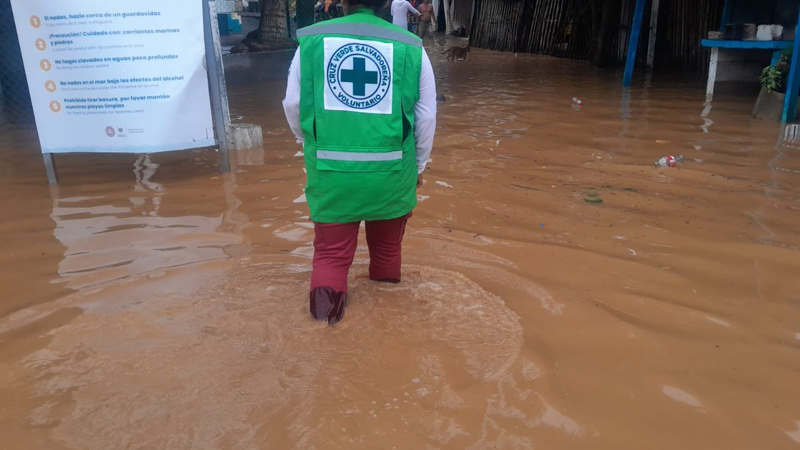 Un voluntario de Cruz Verde Salvadoreña, con su chaleco reflectante, camina con el agua a la rodilla en una calle inundada cerca de la costa. A la izquierda, un cartel de advertencia de la playa sigue en pie. La imagen muestra la rápida evaluación de daños y el inicio de la asistencia en zonas vulnerables.