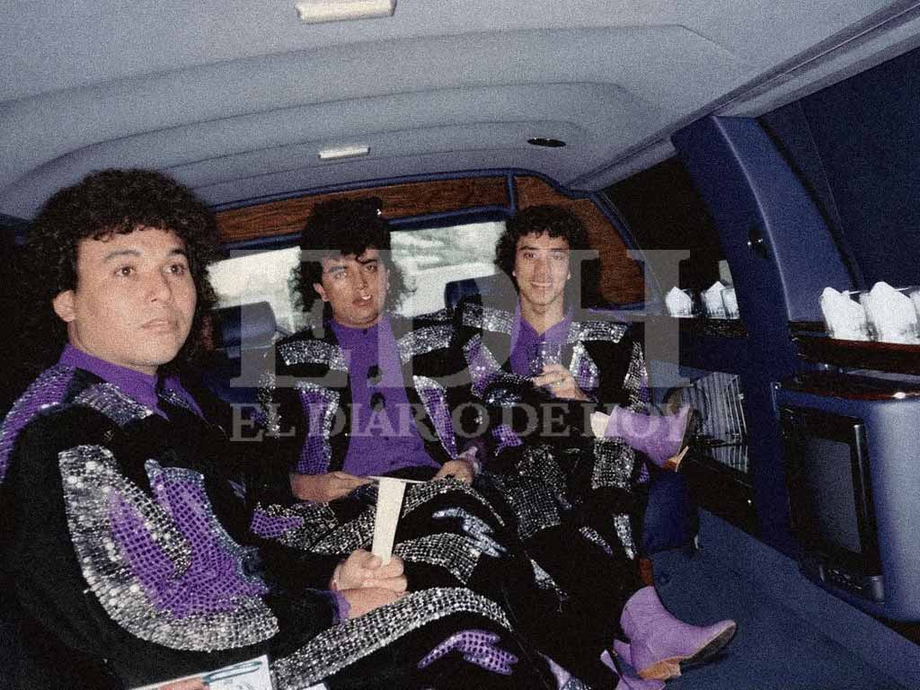 Tres integrantes del Grupo Algodón sentados dentro de una limusina, vistiendo trajes negros brillantes con lentejuelas moradas y plateadas.