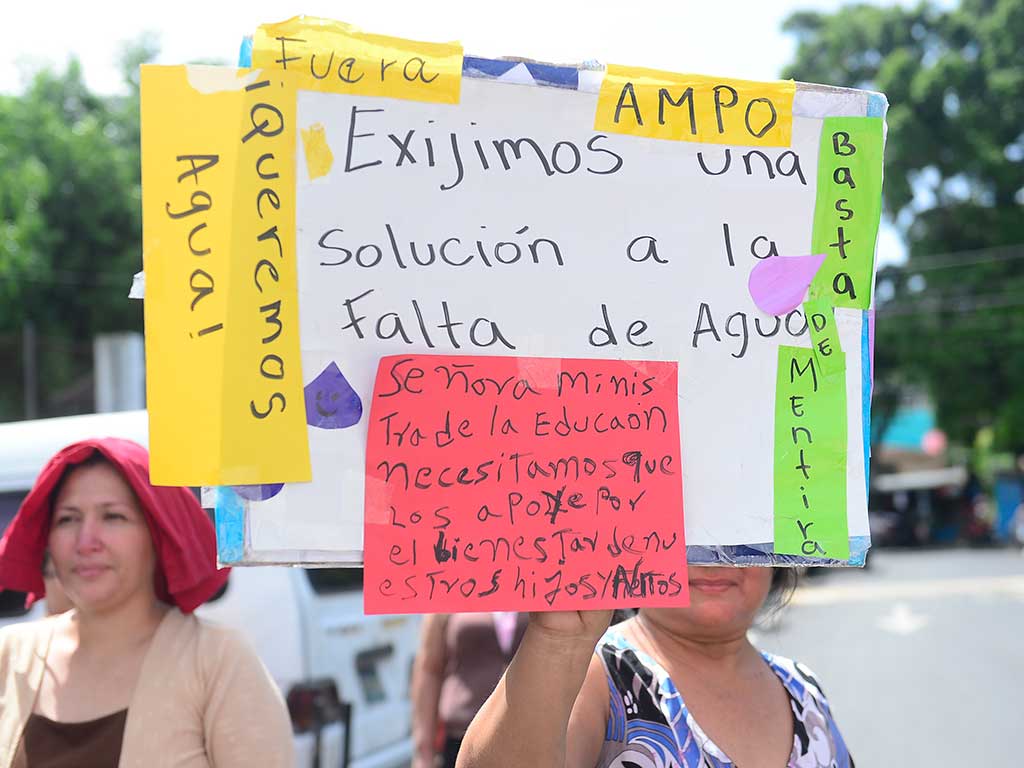Primer plano de una pancarta escrita a mano sobre cartones amarillos y blancos. En rojo dice: "Señora Ministra de la Educaón Necesitamos que nos apolle Por el bienestar de nuestros hijos y Nietos". Al lado, "Queremos Agua!".