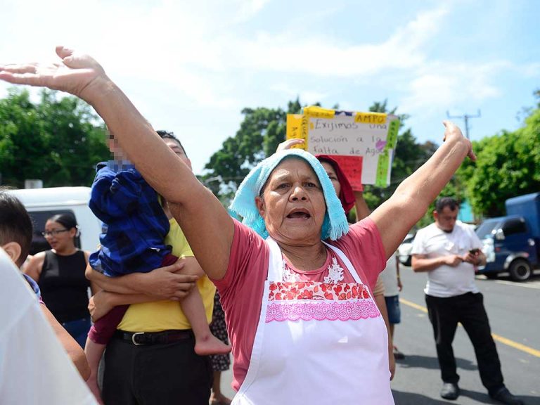 Plano medio de una mujer adulta mayor con pañoleta azul turquesa y delantal, alzando ambos brazos con las manos abiertas hacia el cielo, gritando con fuerza. Detrás, personas cargando niños y carteles.