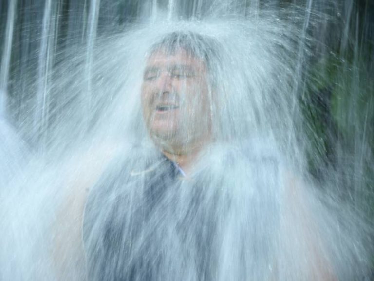 otografía en plano medio de un hombre sonriendo con los ojos cerrados mientras una cortina de agua natural y cristalina cae con fuerza sobre su cabeza en el Turicentro Los Chorros.