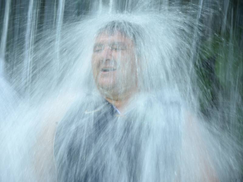 otografía en plano medio de un hombre sonriendo con los ojos cerrados mientras una cortina de agua natural y cristalina cae con fuerza sobre su cabeza en el Turicentro Los Chorros.