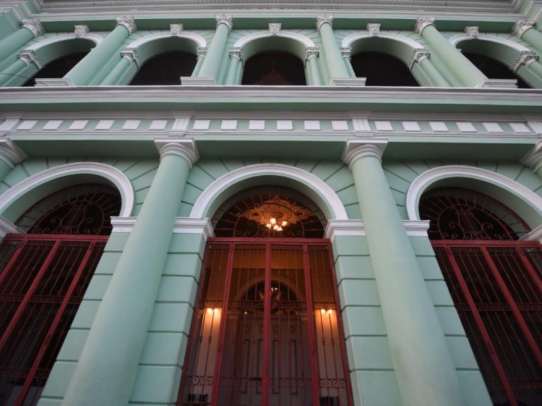 Plano contrapicado cercano de la planta baja del teatro, enfocado en las puertas principales de acceso. Las entradas están protegidas por grandes portones de hierro forjado pintados de rojo oscuro. A través del arco central se observa la luz cálida del interior y un detalle decorativo dorado en el techo del vestíbulo.