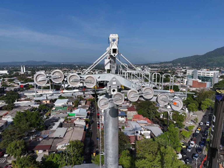 Fotografía a nivel del cabezal de una pilona metálica contra un cielo azul. Se aprecian detalladamente los conjuntos de poleas circulares de metal alineados a ambos lados de la estructura superior. Al fondo a la derecha se observa el brazo de una grúa y abajo se ve una panorámica de la zona urbana con autos en las calles.