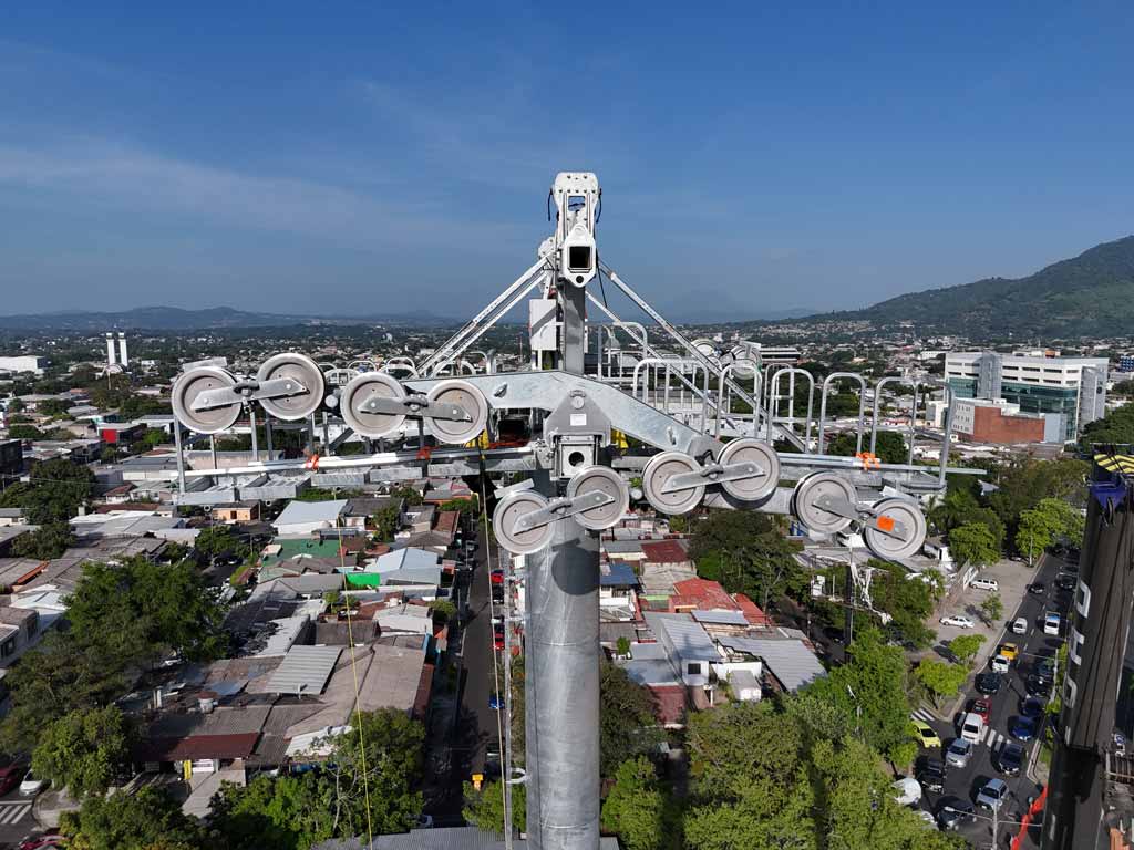 Fotografía a nivel del cabezal de una pilona metálica contra un cielo azul. Se aprecian detalladamente los conjuntos de poleas circulares de metal alineados a ambos lados de la estructura superior. Al fondo a la derecha se observa el brazo de una grúa y abajo se ve una panorámica de la zona urbana con autos en las calles.