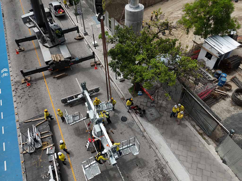Vista aérea que muestra una grúa de construcción y a varios trabajadores con uniformes amarillos en la calle trabajando en la instalación de las estructuras metálicas de la estación del Metrocable. Al fondo se ve la columna de hormigón de la futura torre.