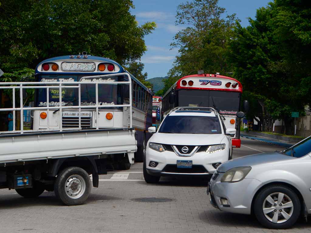 Primer plano del tráfico en la ciudad. Se observa una camioneta blanca Nissan liderando una fila de vehículos, incluyendo dos autobuses coloridos de transporte público, esperando en una intersección de la calle debido a las obras del Metrocable.