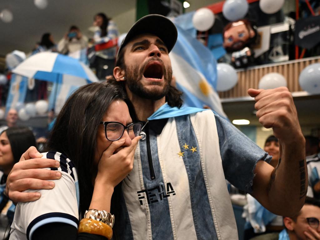 Una imagen que captura la montaña rusa de emociones del fútbol: un hincha con una gorra y una capa de la bandera argentina grita con el alma y el puño apretado, mientras abraza a una mujer que llora de la emoción cubriéndose el rostro con la mano.