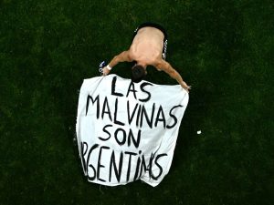 Vista cenital de un jugador argentino (o un hincha que ingresó al campo) arrodillado sobre el césped, sosteniendo con los brazos abiertos una bandera blanca con la contundente leyenda escrita a mano: "LAS MALVINAS SON ARGENTINAS".