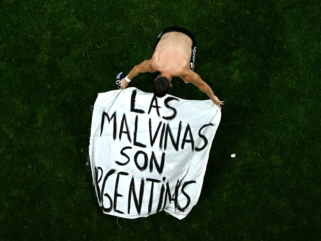 Vista cenital de un jugador argentino (o un hincha que ingresó al campo) arrodillado sobre el césped, sosteniendo con los brazos abiertos una bandera blanca con la contundente leyenda escrita a mano: "LAS MALVINAS SON ARGENTINAS".