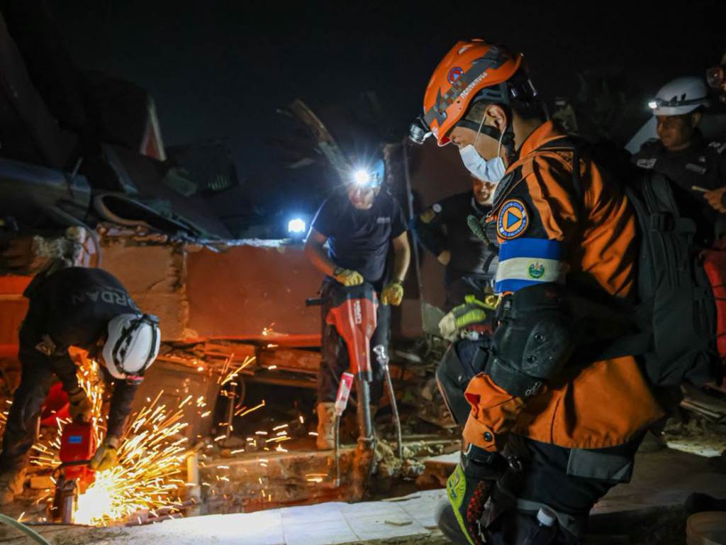 Rescatistas trabajando de noche entre los escombros de una estructura colapsada; se aprecian chispas de corte de metal y herramientas de perforación en movimiento.
