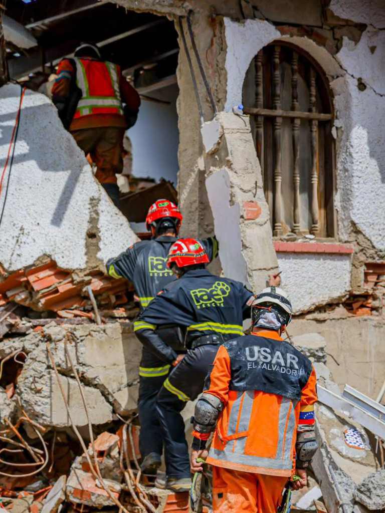 Miembros del equipo de rescate USAR El Salvador ingresando de espaldas y con precaución a una edificación blanca severamente dañada y agrietada.