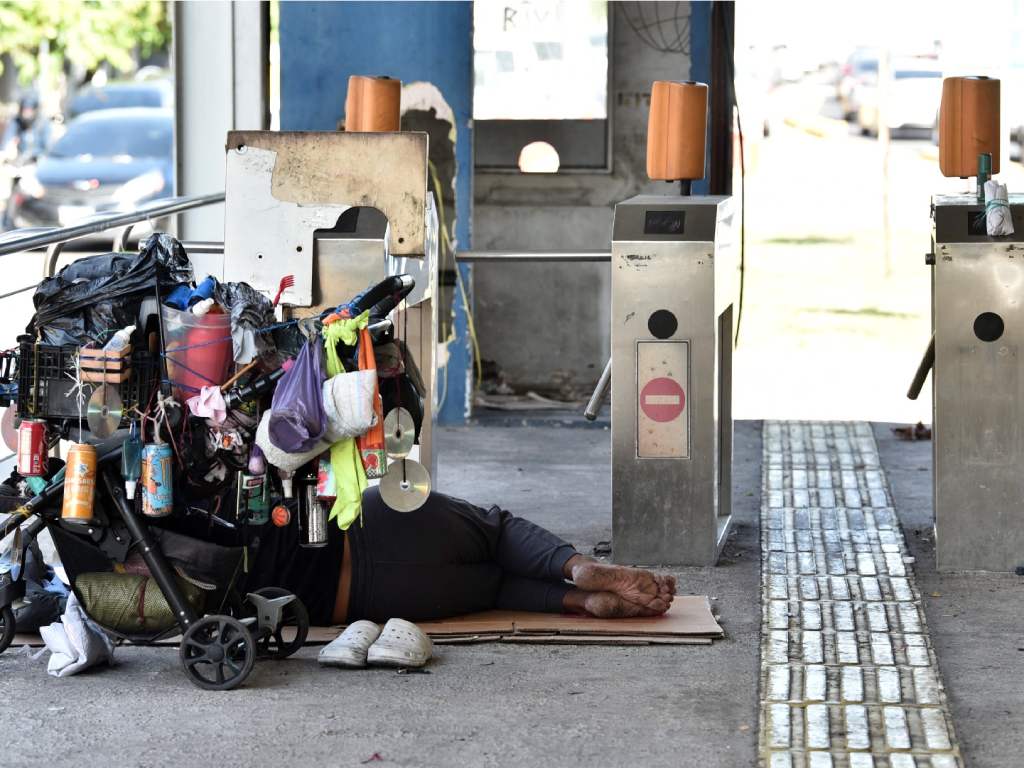 En primer plano, un hombre descansa recostado sobre un colchón deteriorado en el suelo, cubierto con una manta gris. Al fondo, otro hombre permanece sentado junto a un equipaje azul. Las columnas metálicas muestran óxido severo y falta de pintura.