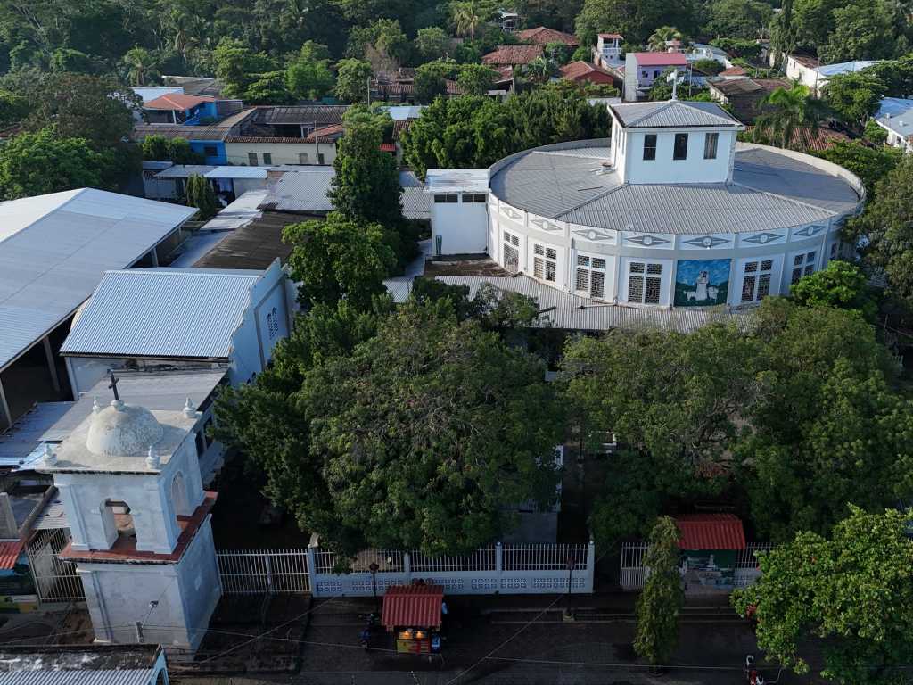 Vista aérea que permite apreciar el templo en su entorno urbano, rodeado de vegetación y las casas de Santiago Nonualco. Desde este ángulo resalta la linternilla o torre central sobre el techo redondo, así como los grandes ventanales con vitrales que bordean la estructura circular del santuario.