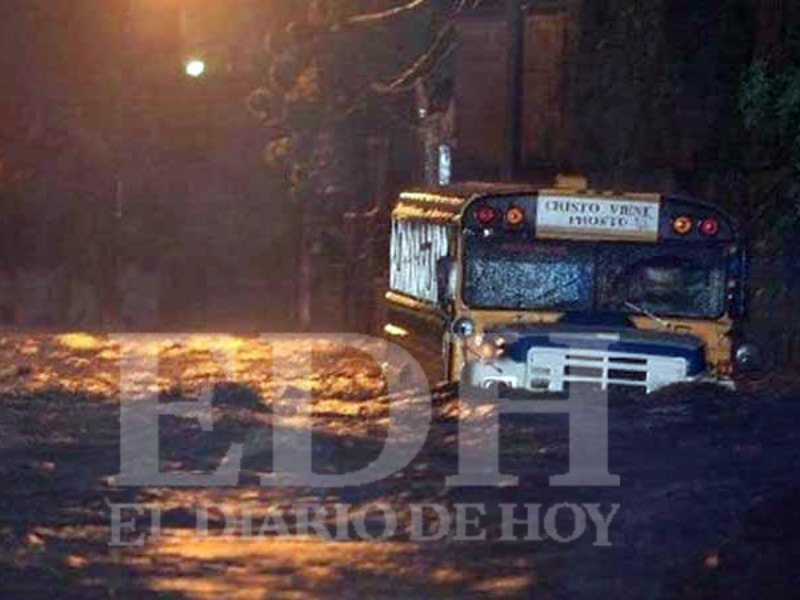 Fotografía nocturna que muestra un autobús escolar amarillo atrapado en medio de una fuerte corriente de agua inundada en una calle. En el letrero frontal del autobús se lee la frase "CRISTO VIENE PRONTO".