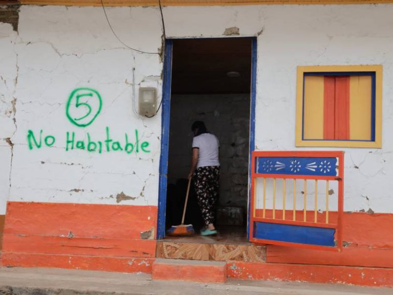 Una mujer sostiene una escoba limpiando el suelo del interior de una vivienda con grietas severas en las paredes y daños estructurales evidentes en el techo tras un terremoto.