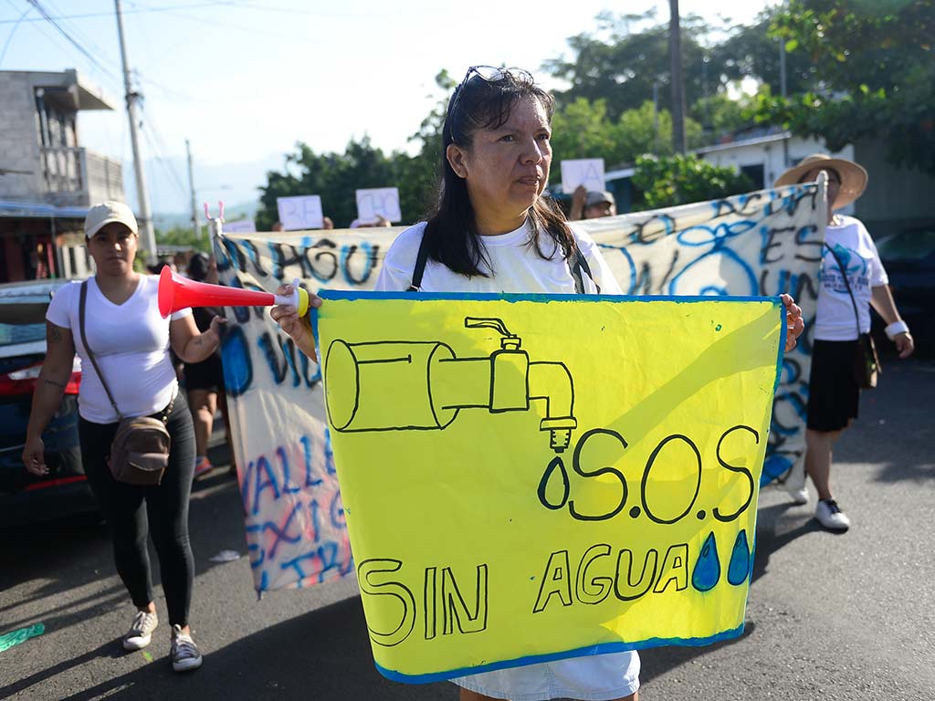 Mujer marcha por la calle sosteniendo una pancarta amarilla dibujada a mano que dice "S.O.S. Sin agua" con el dibujo de un grifo, acompañada por otra mujer con un altavoz de juguete.