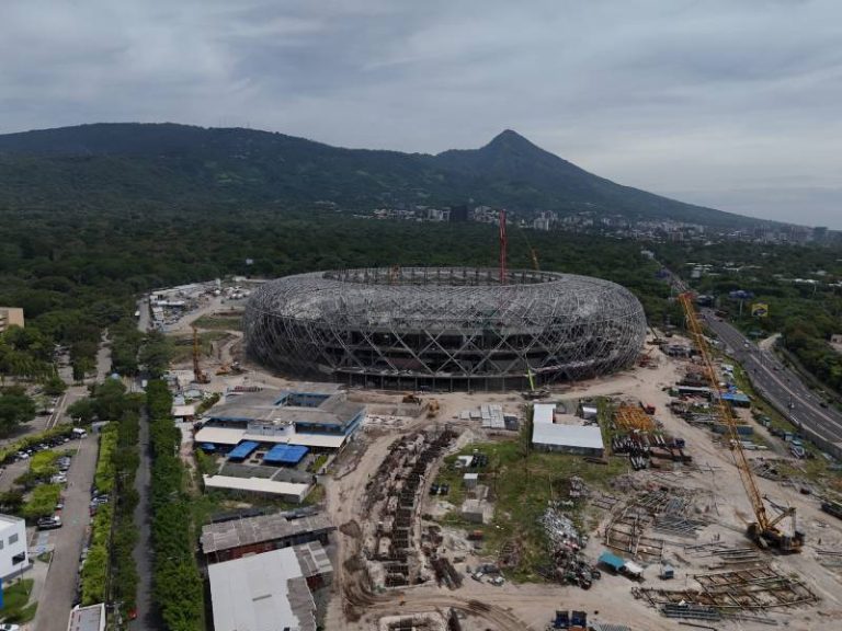 Panorama aéreo del Estadio Nacional en construcción con su armazón metálico circular, grúas en el terreno y un volcán al fondo.