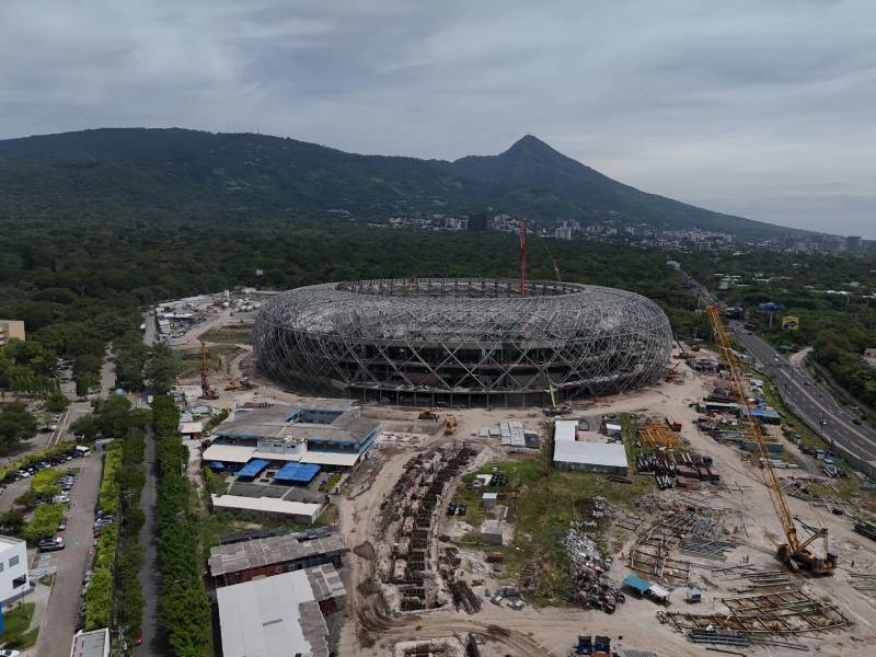 Panorama aéreo del Estadio Nacional en construcción con su armazón metálico circular, grúas en el terreno y un volcán al fondo.