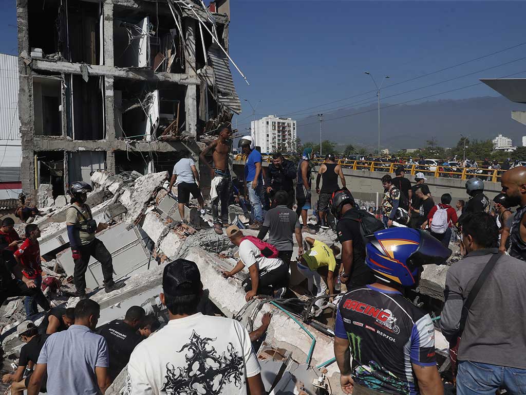 Una multitud de personas, incluyendo socorristas con chalecos y civiles, se agolpa sobre un enorme cúmulo de escombros de concreto y metal frente a un edificio de varios pisos parcialmente derrumbado. El cielo está despejado.