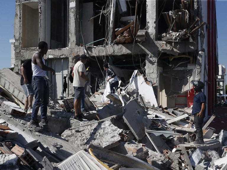 Varios hombres están de pie sobre una gran pila de escombros frente a la fachada expuesta de un edificio de varios pisos que ha perdido sus paredes exteriores. Los pisos están llenos de cables y tuberías colgantes.