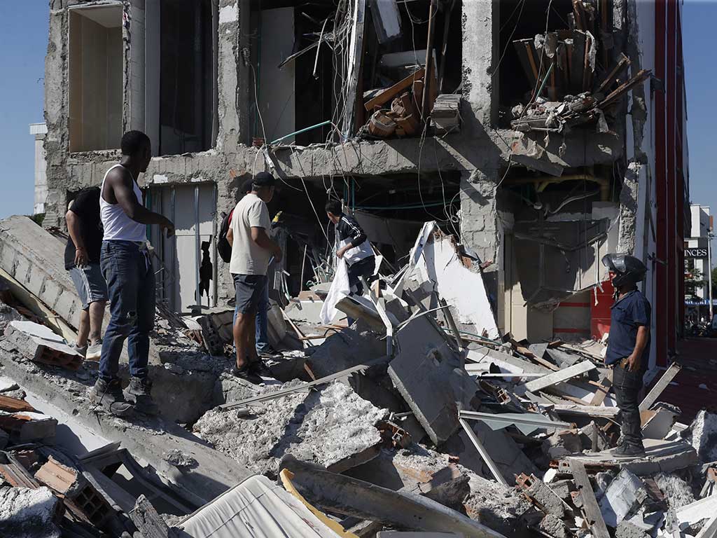 Varios hombres están de pie sobre una gran pila de escombros frente a la fachada expuesta de un edificio de varios pisos que ha perdido sus paredes exteriores. Los pisos están llenos de cables y tuberías colgantes.