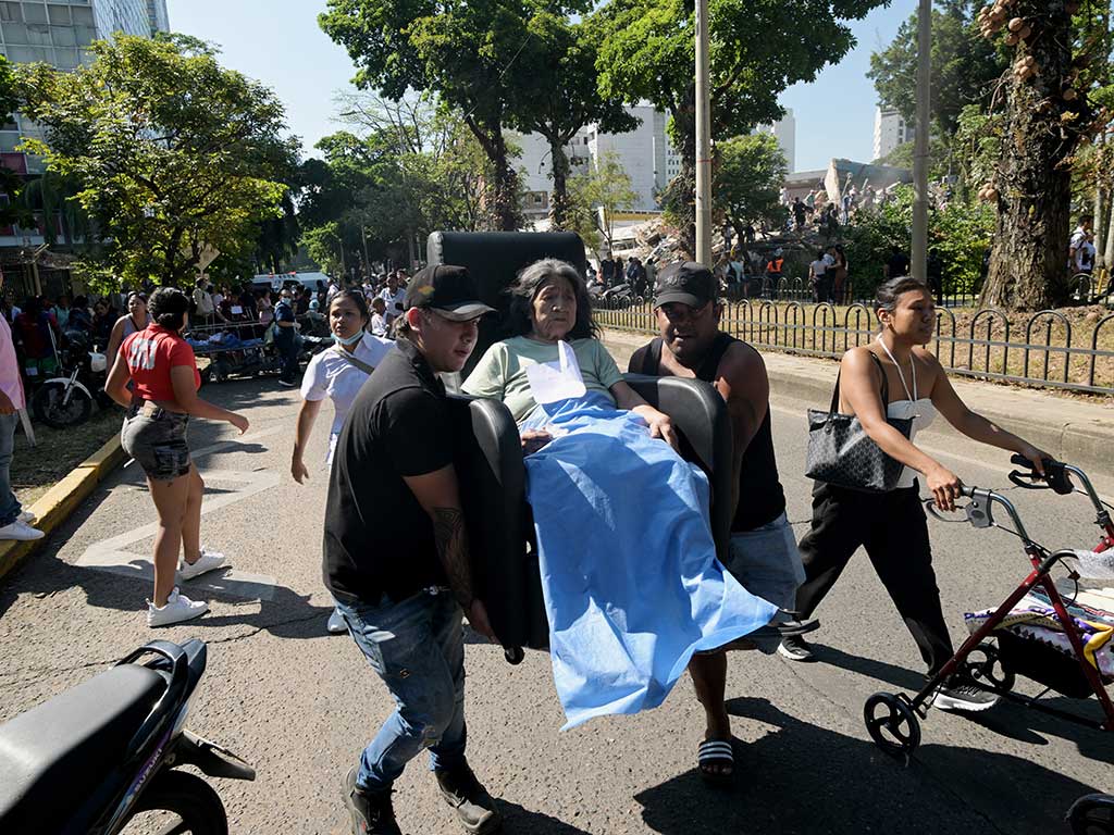 Un grupo de personas carga a una anciana en una silla de oficina por una calle llena de gente. La mujer mayor está cubierta con una sábana azul. Al fondo, un edificio derrumbado y árboles.