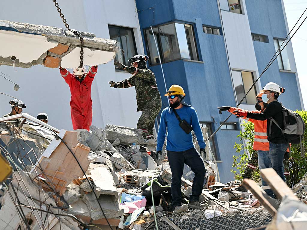 Equipos de socorro y soldados coordinando el levantamiento de una gran losa de concreto sostenida por cadenas sobre un edificio derrumbado.