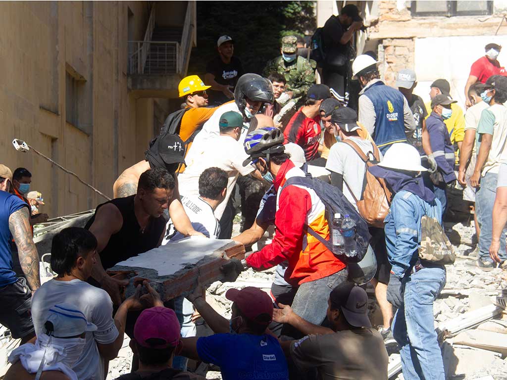 Grupo denso de hombres cargando y trasladando manualmente un pedazo grande de pared de ladrillos entre las ruinas.
