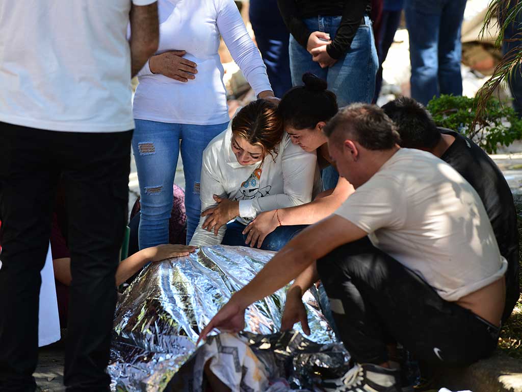 Personas agrupadas e inclinadas en el suelo llorando junto a un cuerpo cubierto con una manta térmica plateada.