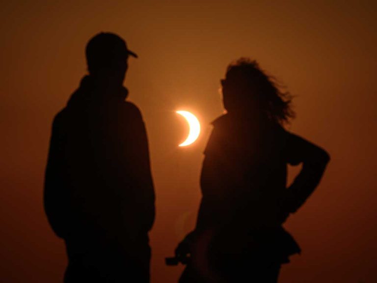 Vista de silueta de dos personas, un hombre con gorra a la izquierda y una mujer con cabello al viento a la derecha, de pie y mirando hacia un eclipse solar parcial en forma de media luna brillante en un cielo teñido de naranja intenso al atardecer.