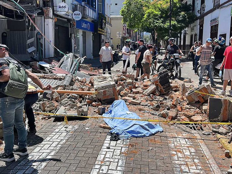 Un cuerpo sin vida cubierto con una sábana azul yace en una calle adoquinada rodeada de escombros de ladrillos, con una cinta amarilla de precaución y peatones observando.
