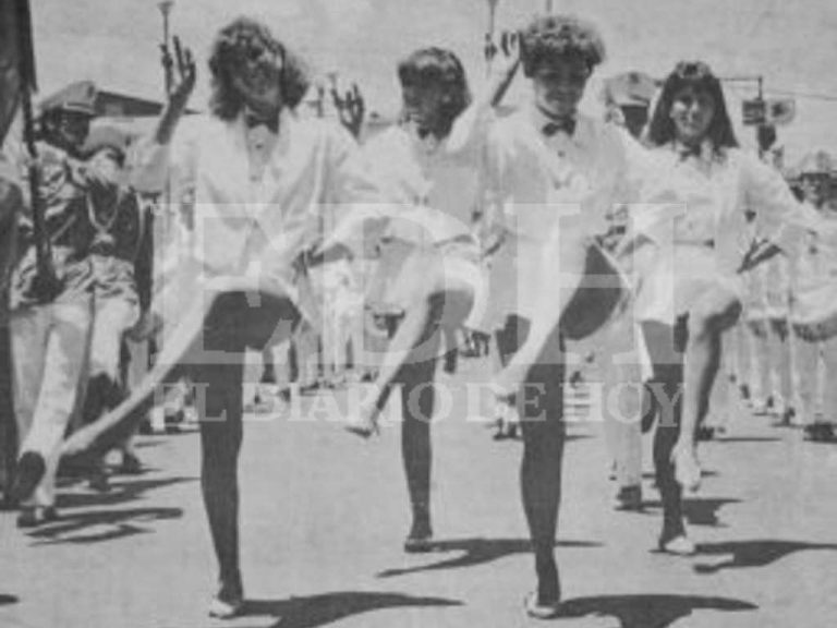 Fotografía en blanco y negro que muestra a cuatro cachiporras ejecutando una coreografía coordinada levantando la pierna durante un desfile militar o escolar en la vía pública.
