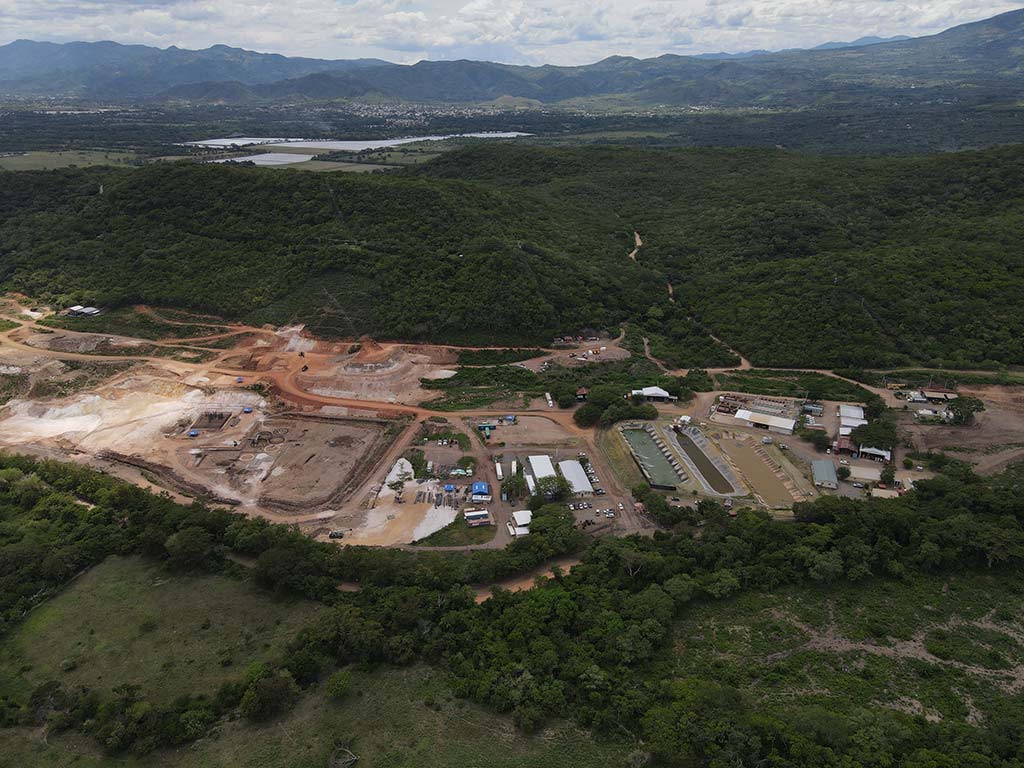 Fotografía aérea en gran angular que muestra un amplio complejo industrial y minero en medio de un denso bosque verde. A la izquierda se observa una gran área descapotada de tierra clara y rojiza con terrazas excavadas; al centro y derecha, instalaciones con naves industriales, contenedores, estanques rectangulares de almacenamiento de agua y caminos de tierra. Al fondo se extienden montañas y un cuerpo de agua bajo un cielo nublado.