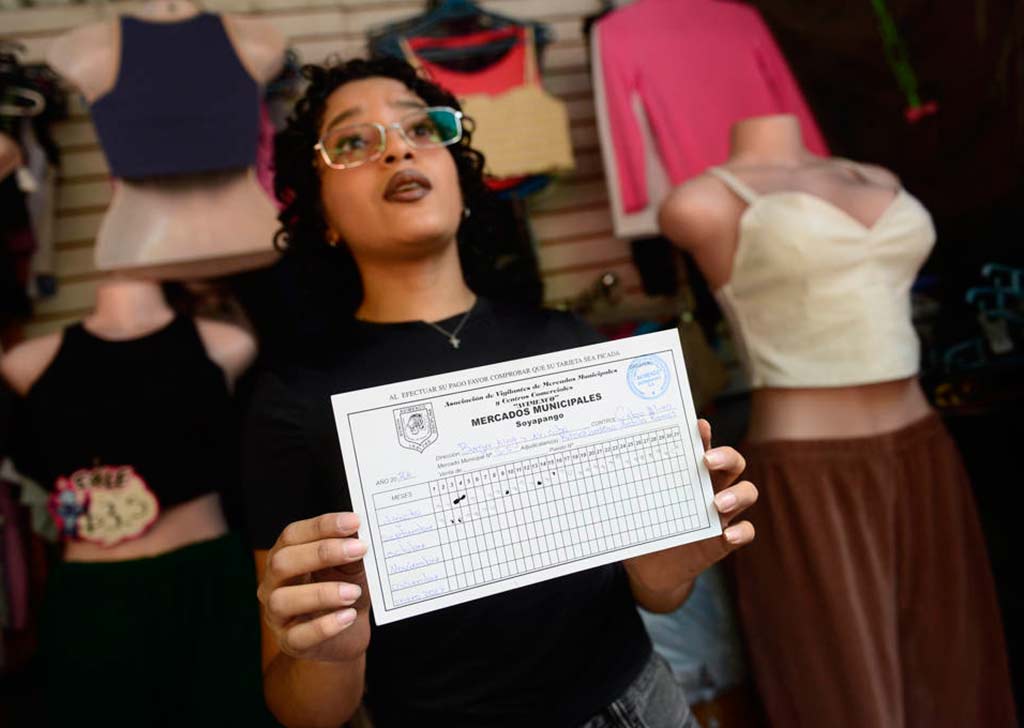Plano medio de una mujer con gafas y cabello oscuro rizado que mira a la cámara mientras sostiene una tarjeta de papel blanco con ambas manos. El texto legible en la tarjeta dice "TARJETA DE CONTROL DE COBROS DIARIOS". El fondo muestra un puesto de ropa desenfocado, con maniquíes y estantes.