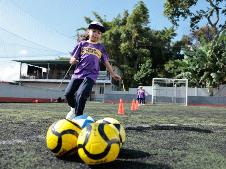 Grupo de niñas y adolescentes jugando al fútbol en una cancha deportiva al aire libre en San Salvador.