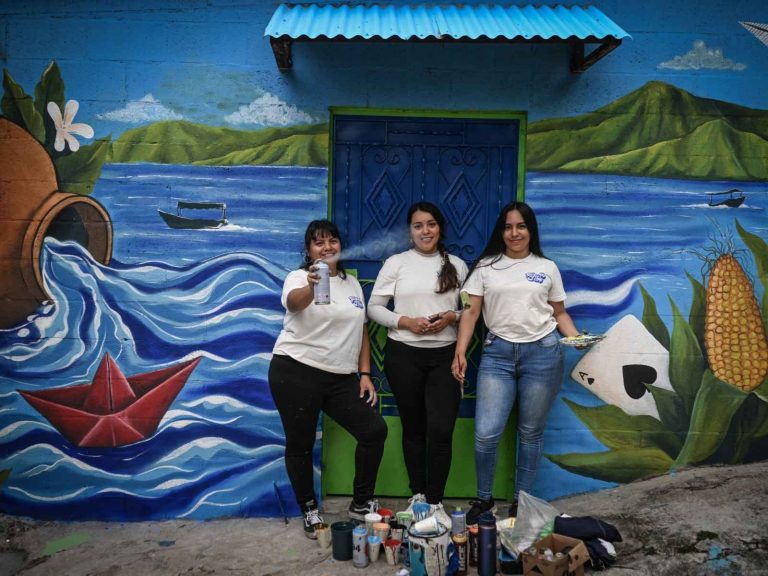Patricia Vega, Marcela Alvarenga y Elizabeth Guardado pintan un mural con aerosoles y pinceles en una comunidad urbana.