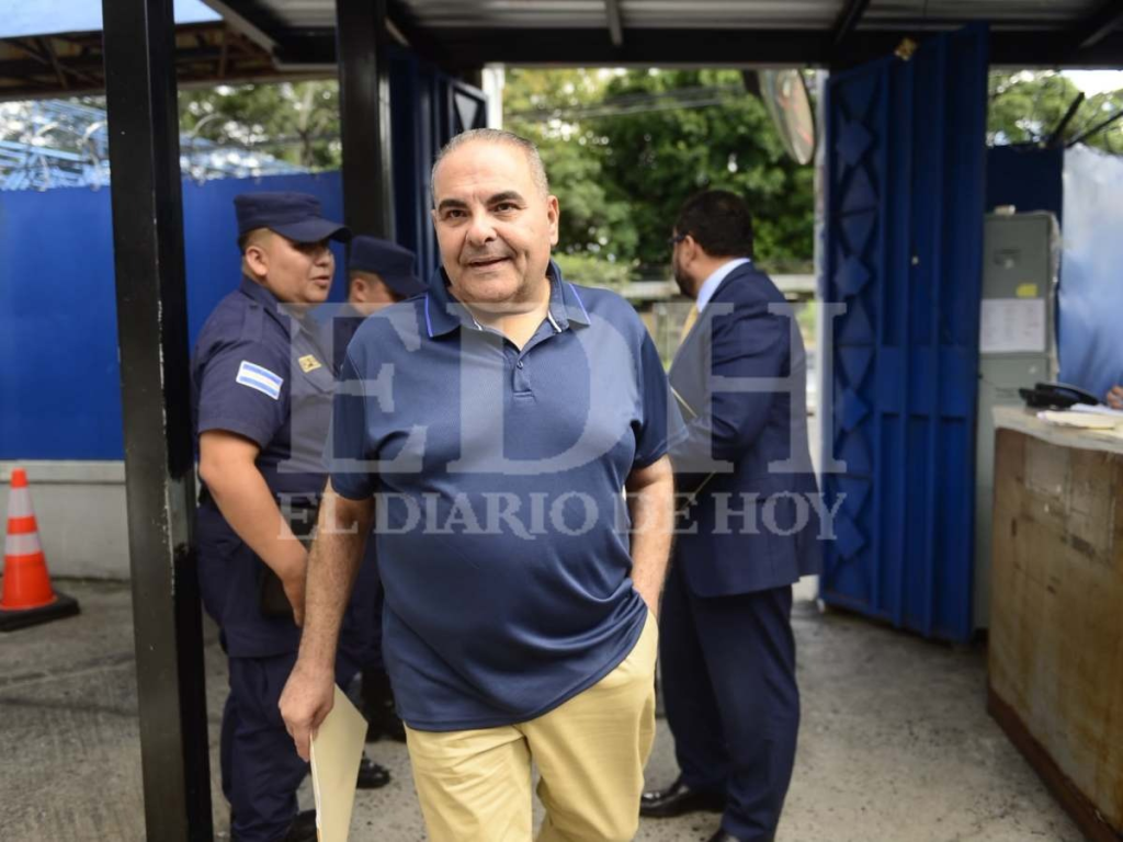 Text Alt: Antonio Saca con camiseta azul polo y pantalón beige caminando al lado de su abogado de traje azul y corbata amarilla en una plaza abierta.