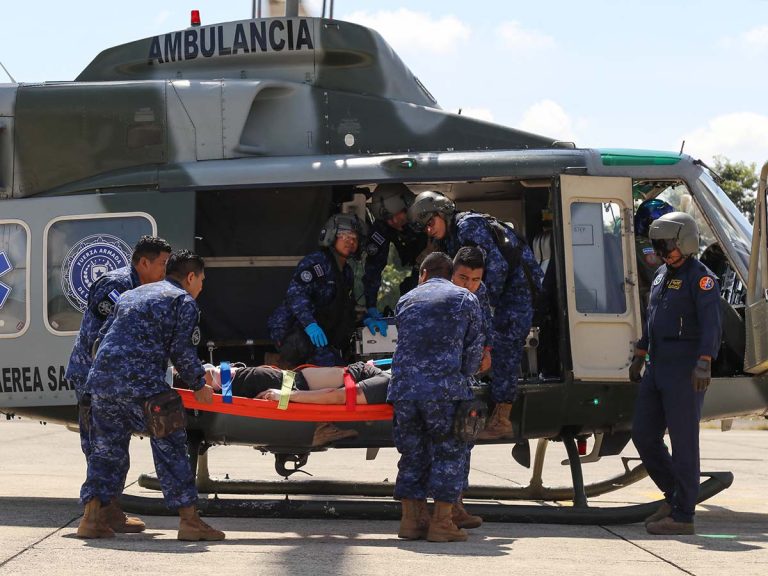 Grupo de militares con uniforme de camuflaje azul sosteniendo una camilla bajo la estructura de un helicóptero militar.